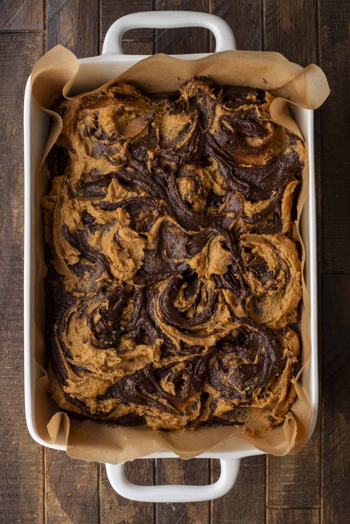 A rectangular baking dish lined with parchment paper holds a batch of swirled brownies, featuring a marbled mix of dark chocolate and lighter, caramel-colored batter. The brownies appear freshly baked and rich.