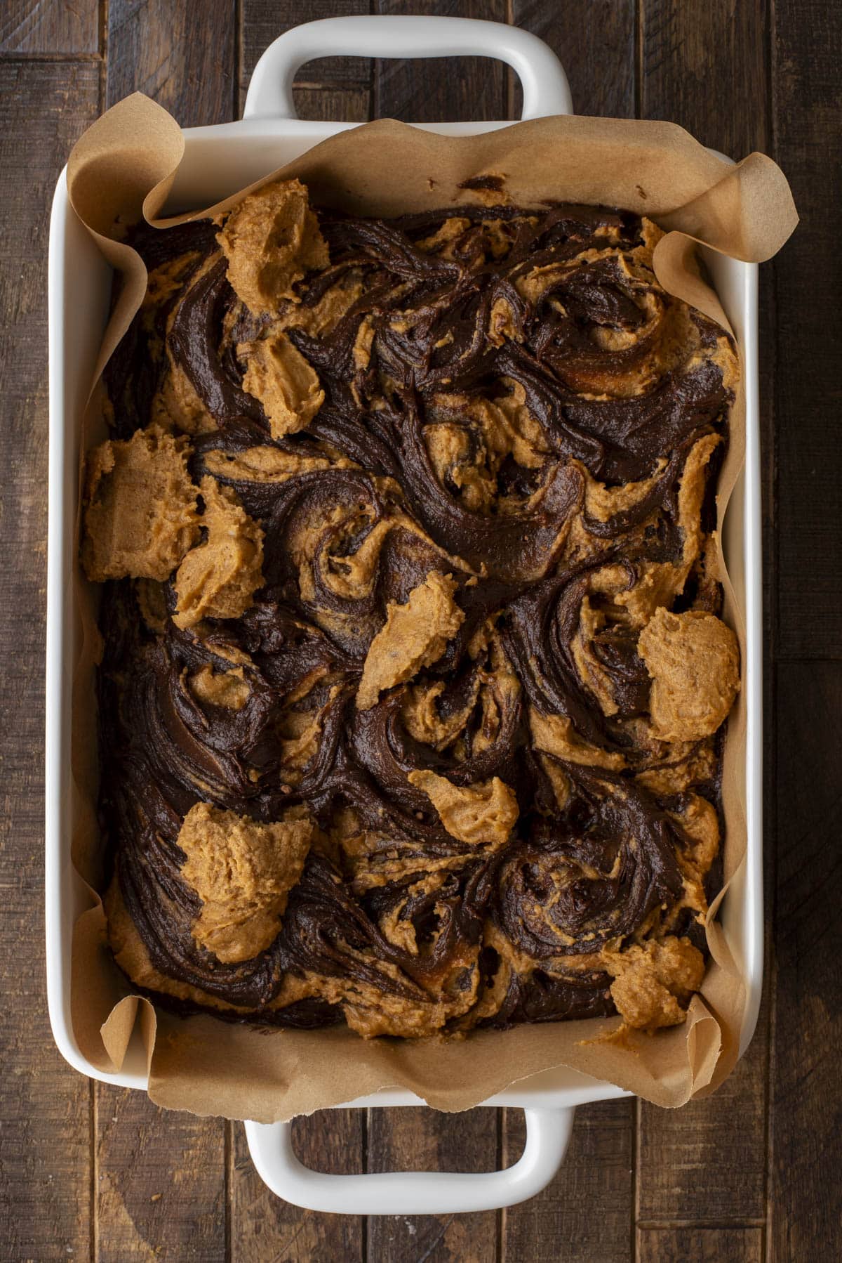 A rectangular baking dish lined with parchment paper holds freshly baked brownies swirled with a light brown peanut butter mixture on top. The dessert has a marbled pattern and rests on a wooden surface.
