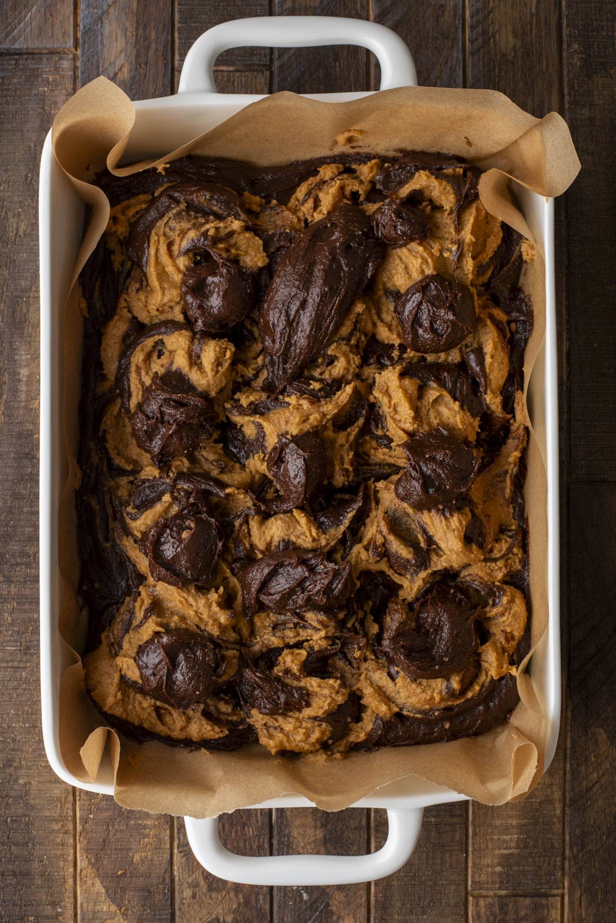A white baking dish lined with parchment paper holds a freshly baked dessert with dark chocolate and golden brown swirls, giving a marbled appearance. The dish is placed on a rustic wooden surface.