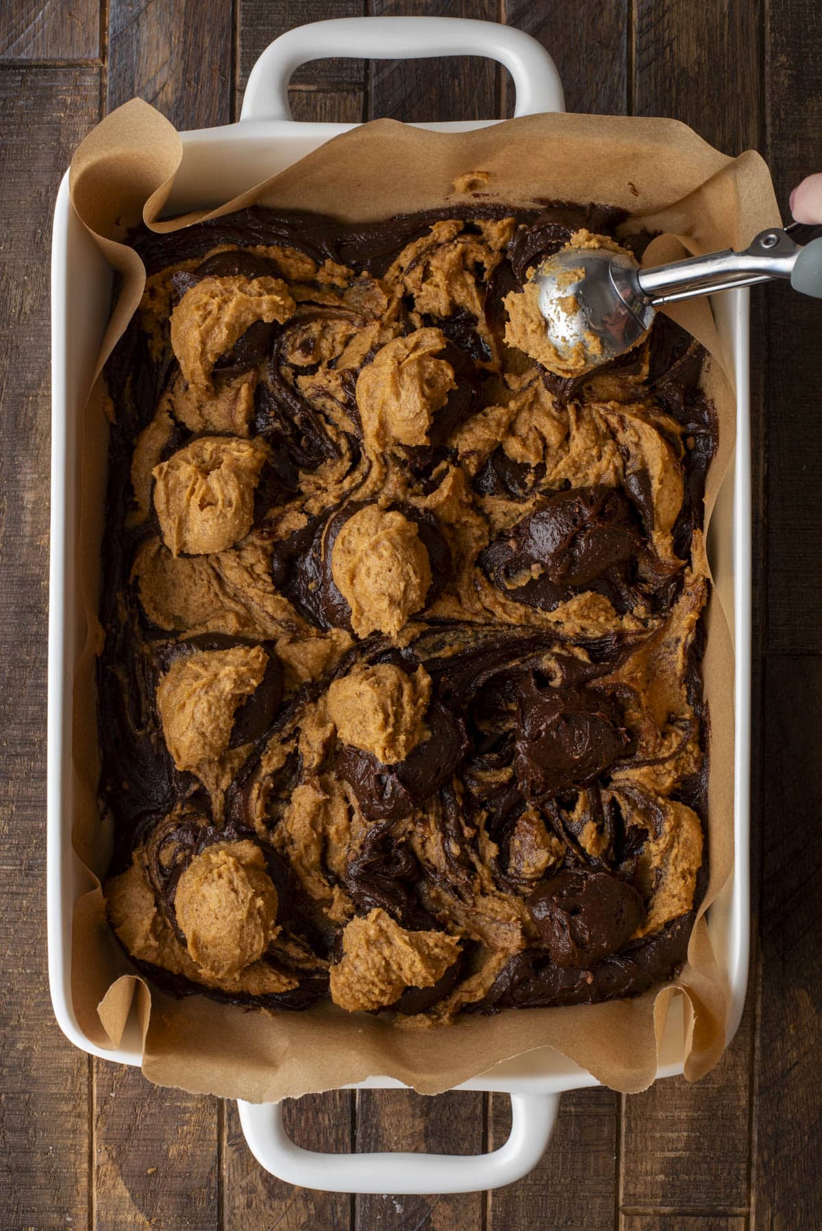 A baking dish lined with parchment paper contains swirled dollops of chocolate and peanut butter brownie batter. A hand is using a cookie scoop to add more batter. The dish rests on a wooden surface.