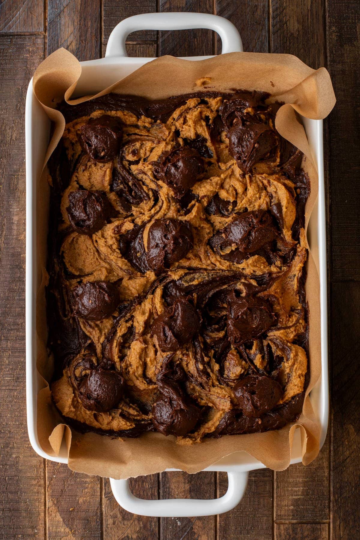 A rectangular baking dish lined with parchment paper holds a batch of chocolate and peanut butter swirled brownies, freshly baked, with rich dark and light swirls visible on top.