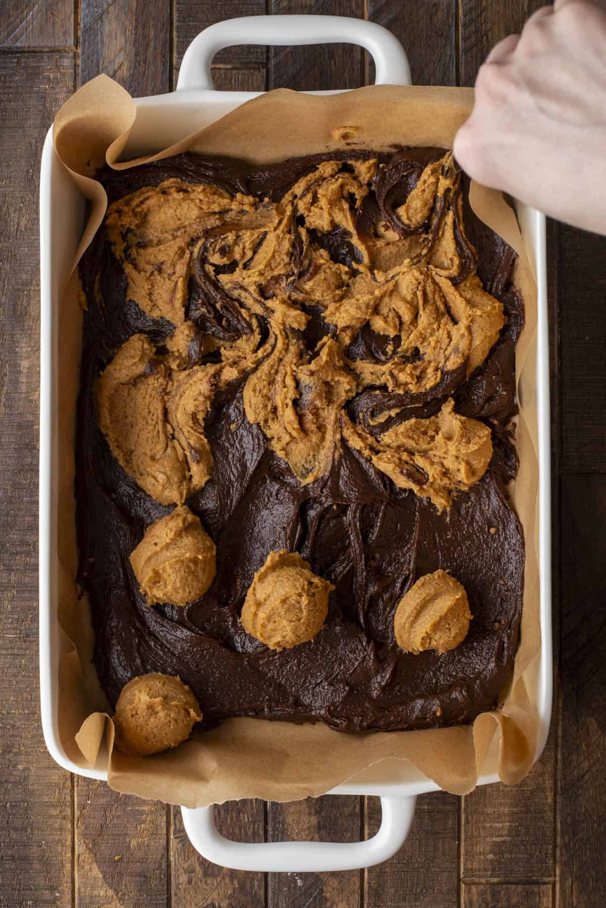 A hand swirls cookie dough into brownie batter in a parchment-lined baking dish, creating a marbled effect. Some scoops of cookie dough are still visible on top of the brownie mixture.