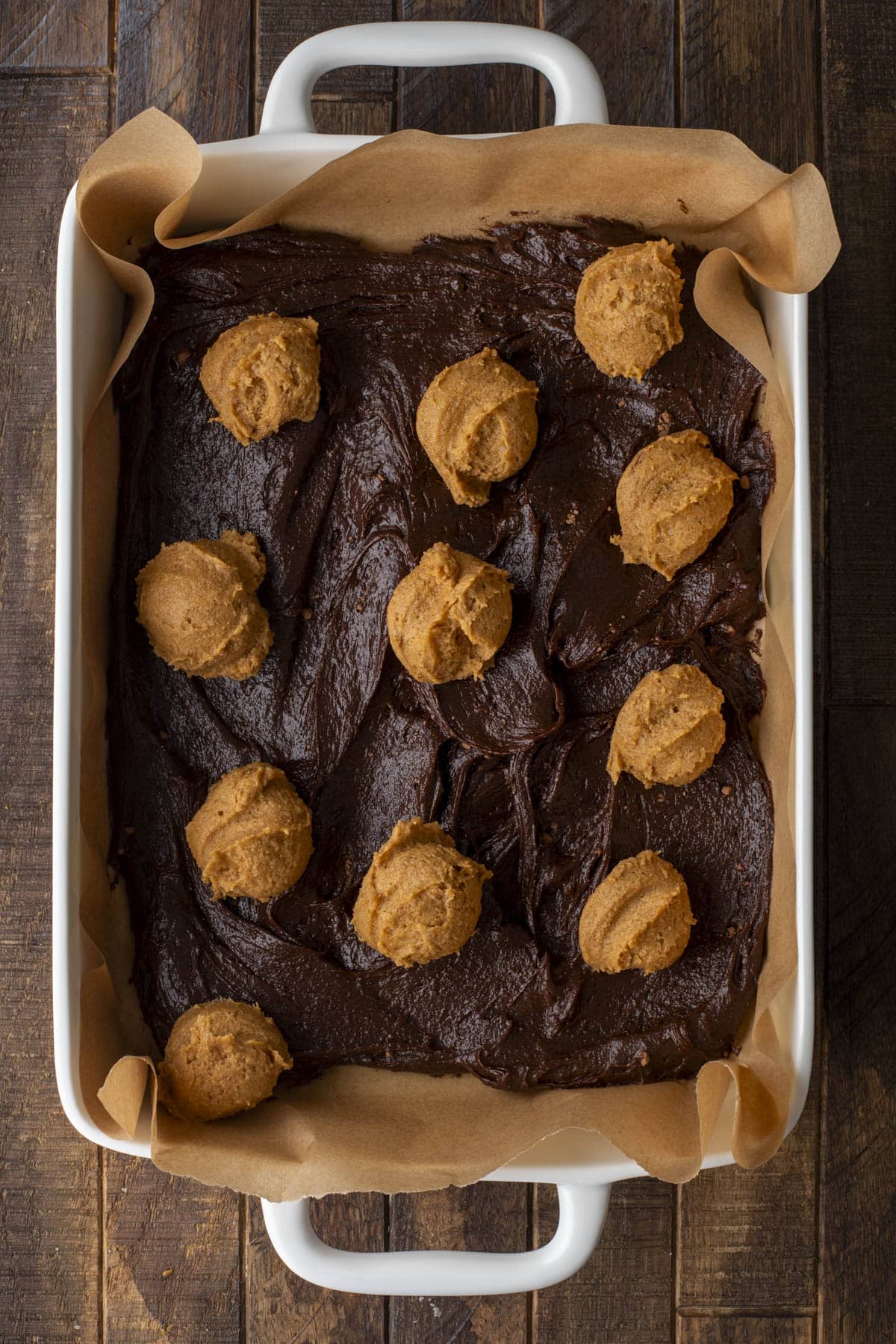 A white baking dish lined with parchment paper contains chocolate brownie batter spread out, with several scoops of peanut butter dough evenly placed on top, ready to be baked.
