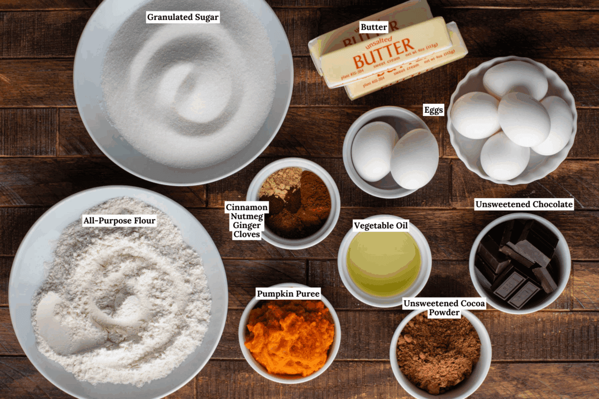 Bowls and containers of labeled baking ingredients—granulated sugar, all-purpose flour, eggs, butter, pumpkin puree, vegetable oil, unsweetened chocolate, spices, and cocoa powder—arranged on a wooden surface.