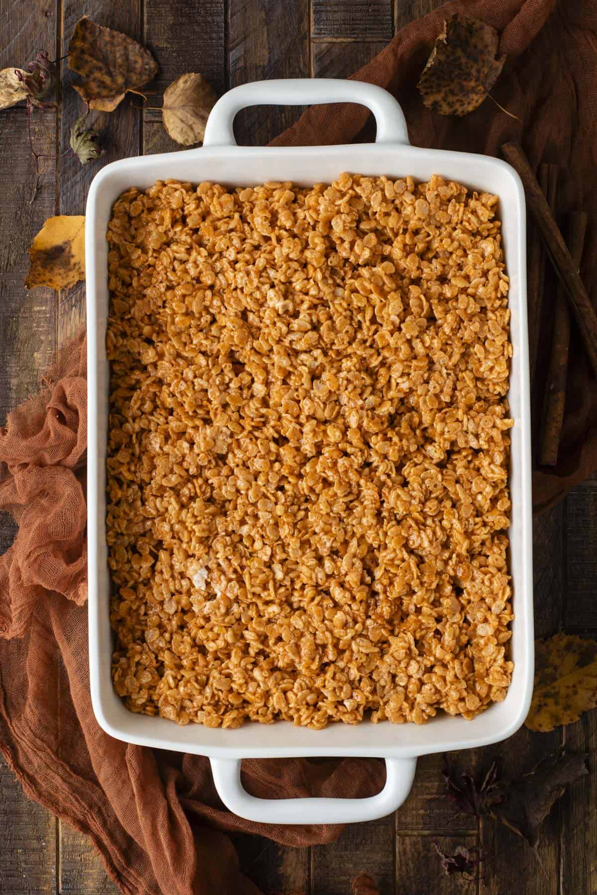 A white rectangular baking dish filled with golden-brown Rice Krispies treats sits on a rustic wooden table, surrounded by brown cloth and autumn leaves.