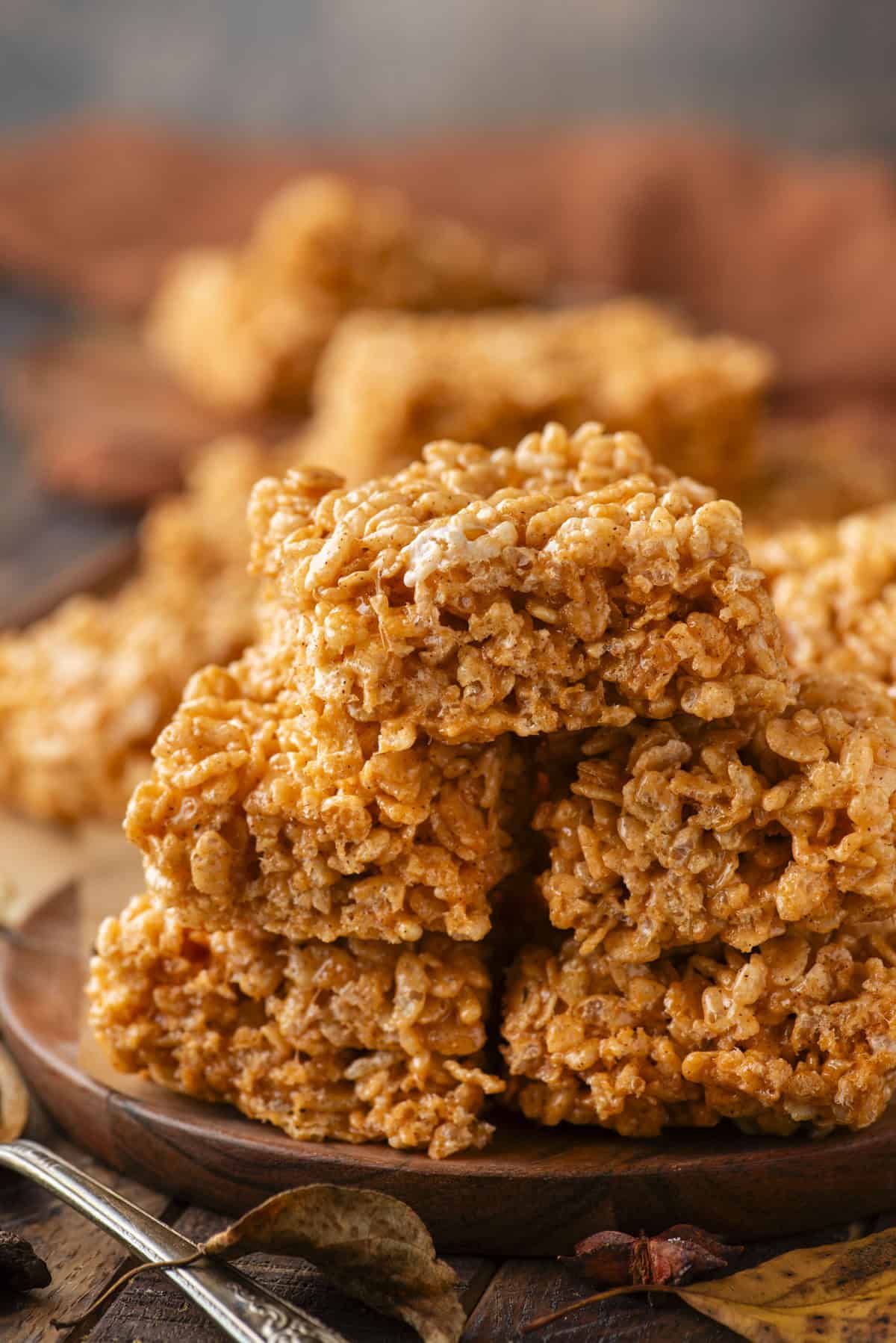 A close-up of a stack of pumpkin spice rice crispy treats on a wooden plate, with more treats and autumn leaves blurred in the background. The treats are golden brown and have a textured, crispy appearance.