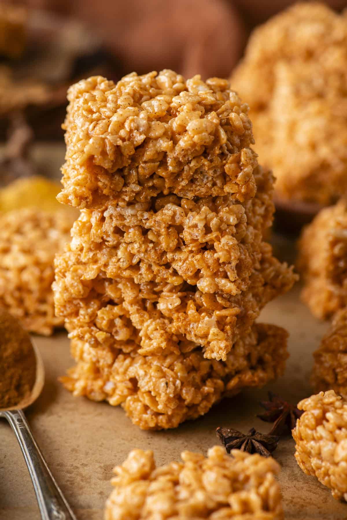 Three pumpkin spice rice crispy treats stacked on top of each other, surrounded by additional treats and a spoon on a brown surface. The treats have a golden orange hue and a textured, crispy appearance.