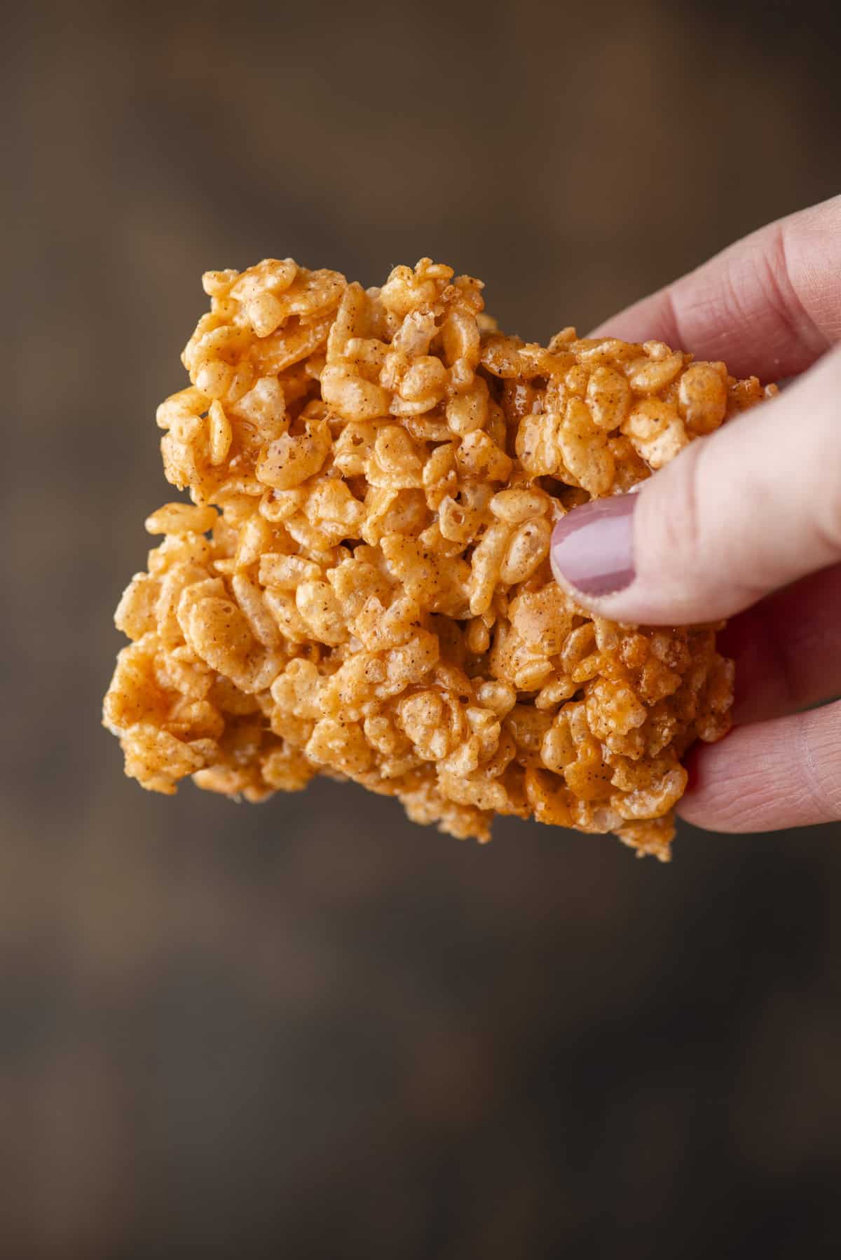 A hand with painted nails holds a single Rice Krispies treat made of puffed rice cereal and marshmallow against a blurred brown background.