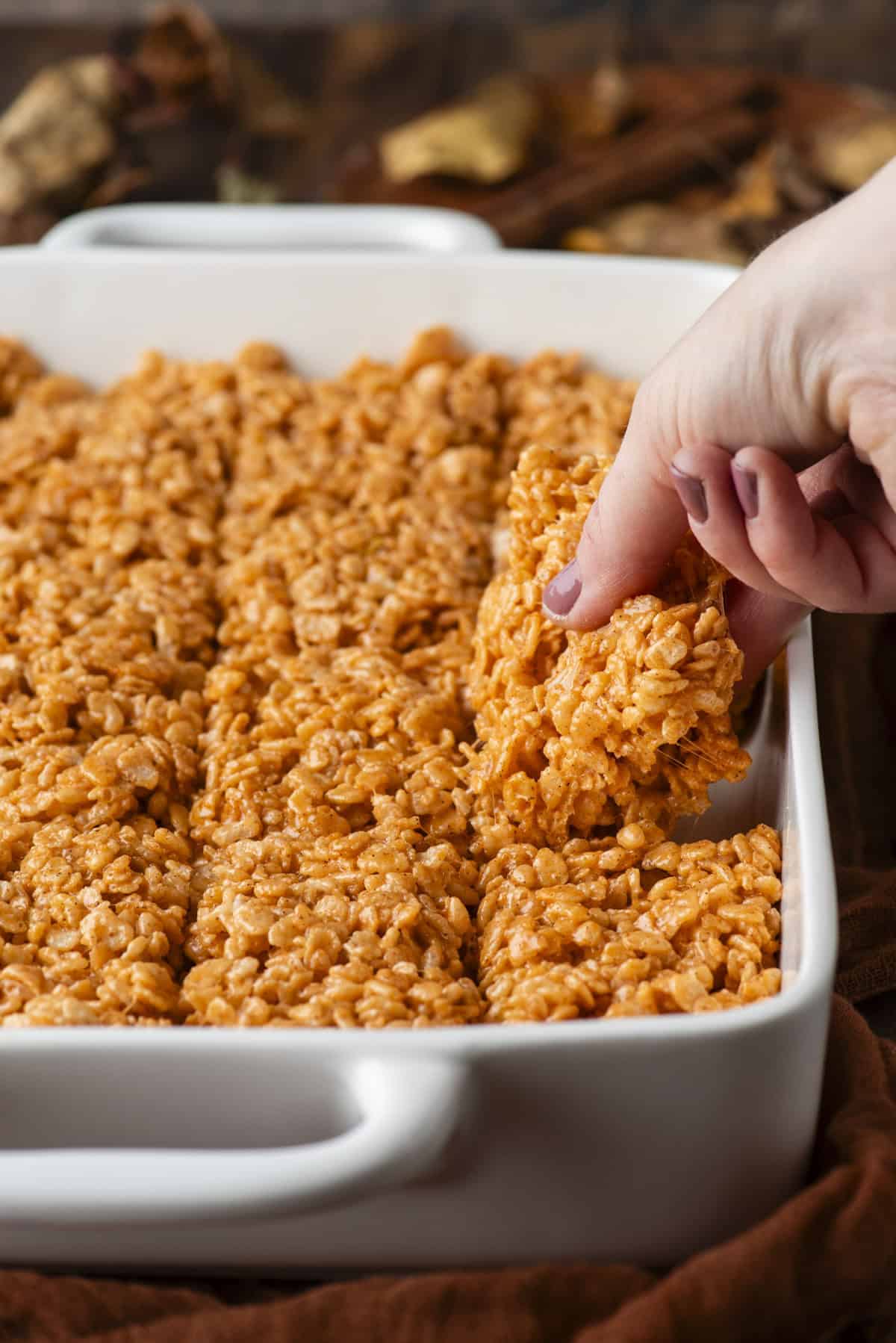 A hand lifts a pumpkin-colored rice crispy treat from a white baking dish filled with neatly cut treats.