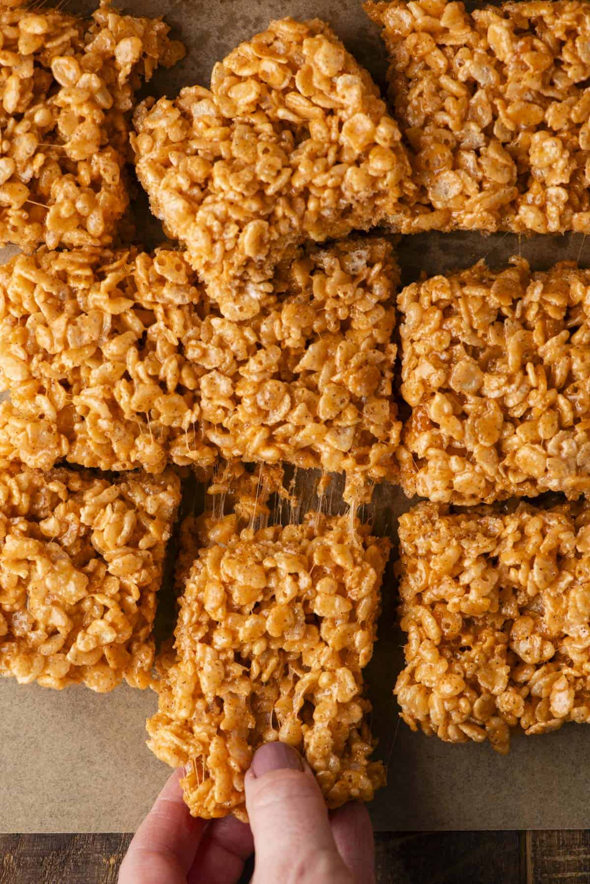 A hand is pulling apart a square of sticky, golden rice crispy treat from a batch of similar squares on a brown surface. The treats have a glossy, marshmallow-coated appearance.