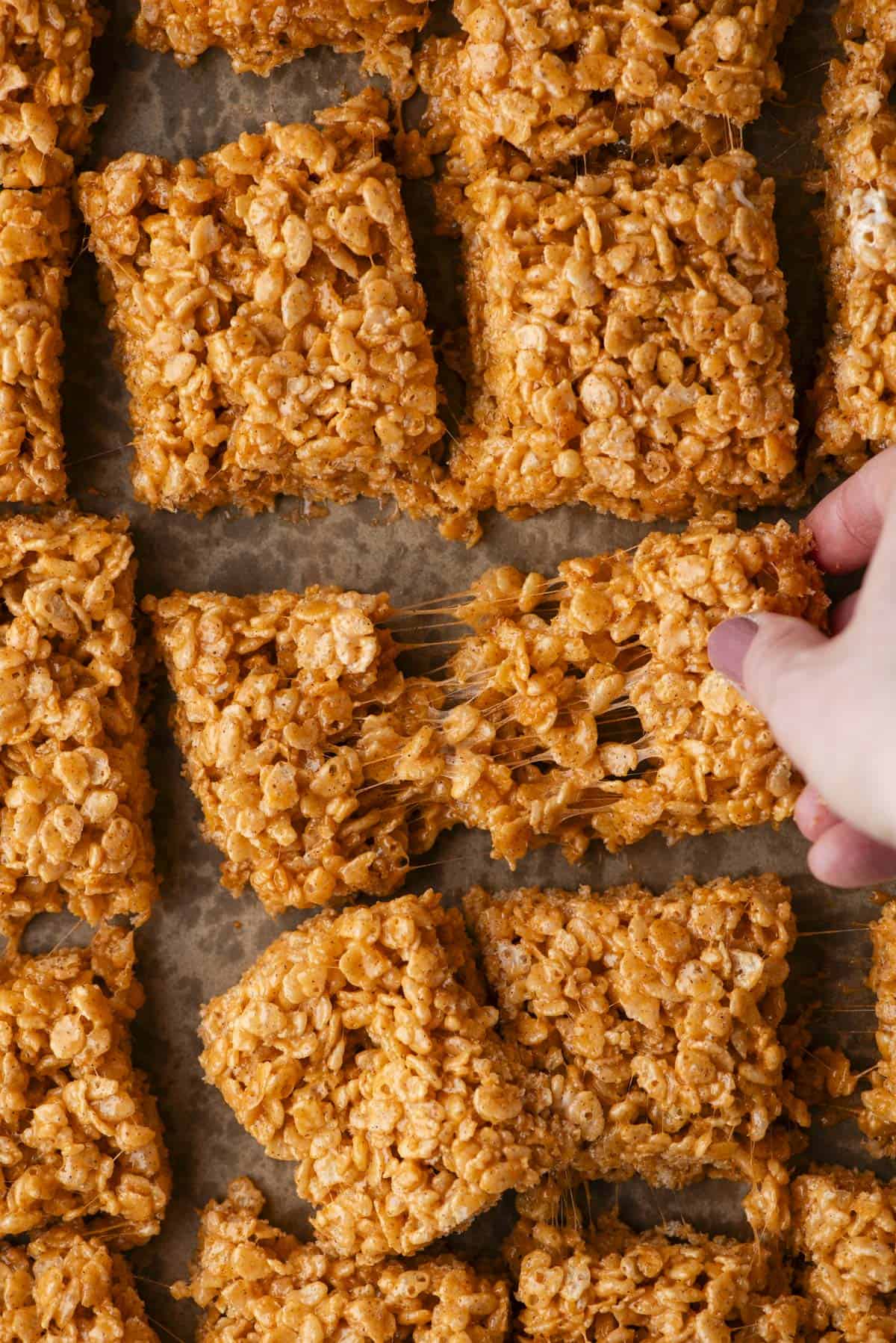 Hand pulling apart a square of golden brown Rice Krispies treat, showing gooey, sticky texture. Several other squares are arranged closely on parchment paper in the background.