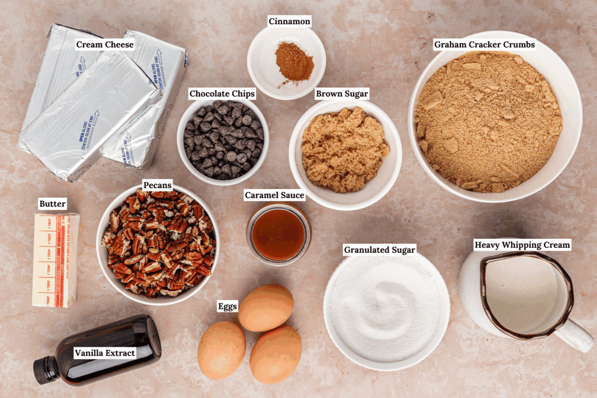 A top-down view of labeled baking ingredients including cream cheese, chocolate chips, pecans, butter, vanilla extract, eggs, cinnamon, caramel sauce, brown sugar, granulated sugar, graham cracker crumbs, and heavy whipping cream.