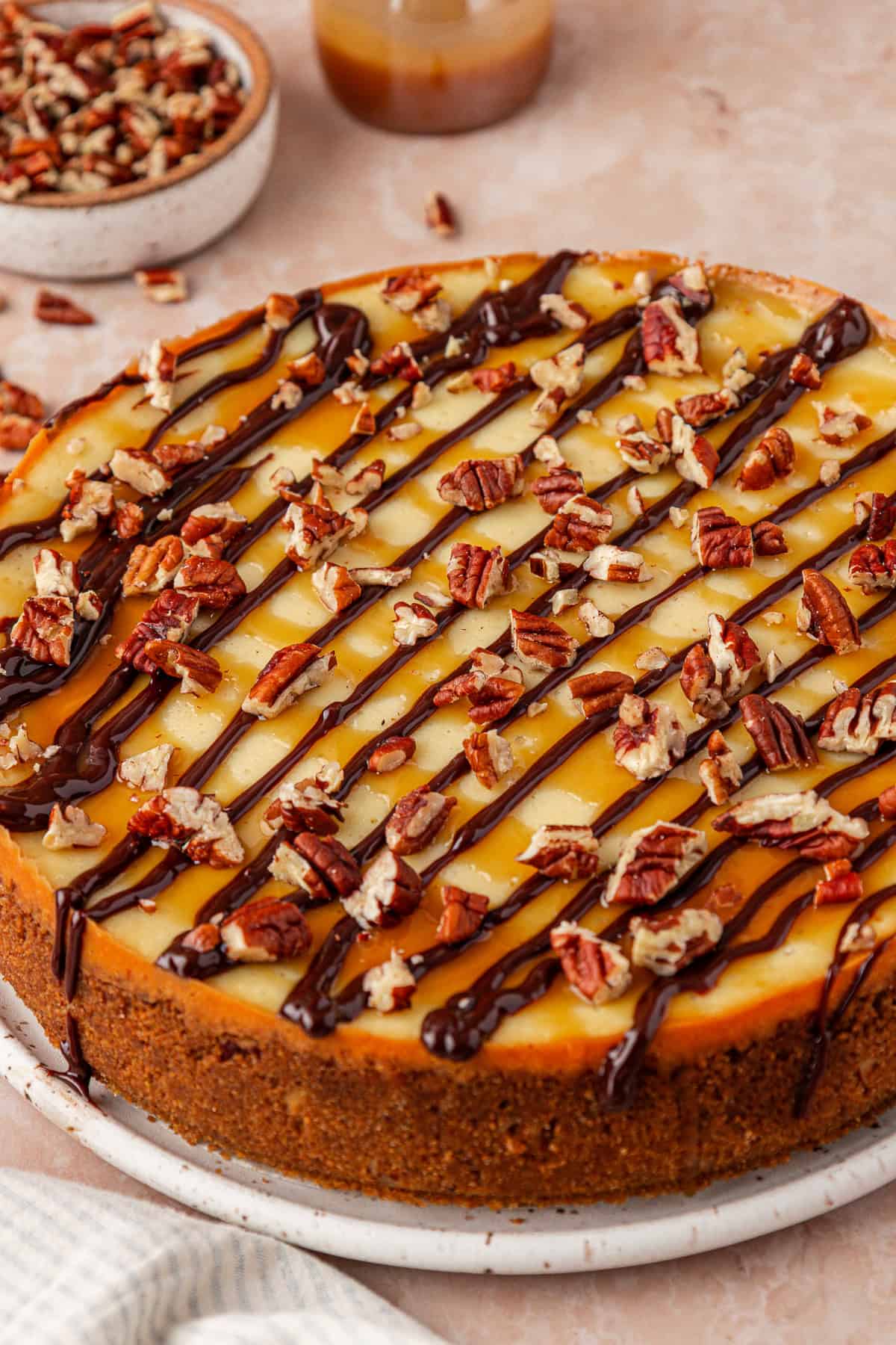 A cheesecake topped with chopped pecans, caramel sauce, and chocolate drizzle, displayed on a round white plate. A bowl of extra pecans and a jar of caramel sauce are in the background.