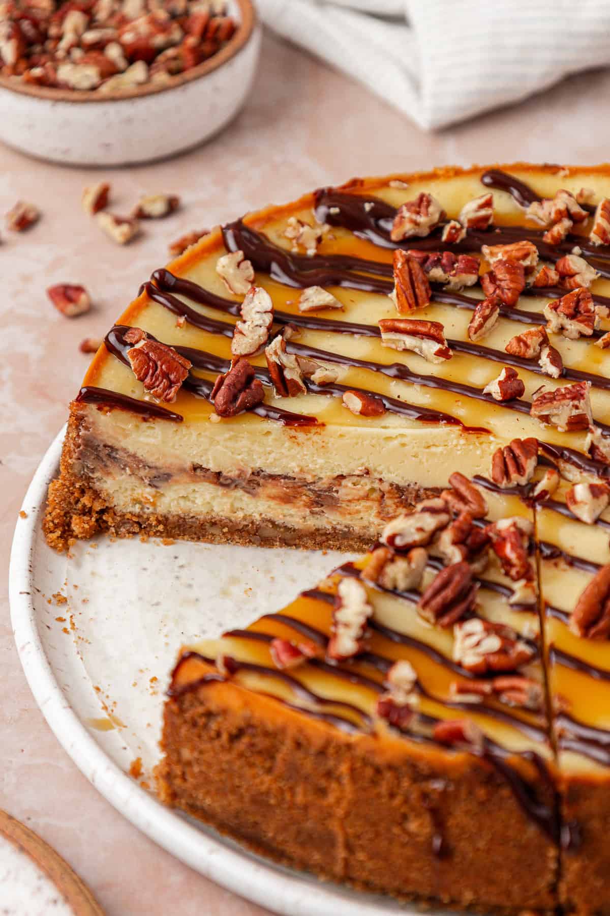 A caramel and chocolate-drizzled cheesecake topped with chopped pecans sits on a white plate, with one slice removed. Crumbs and extra pecans are scattered around the plate and table.