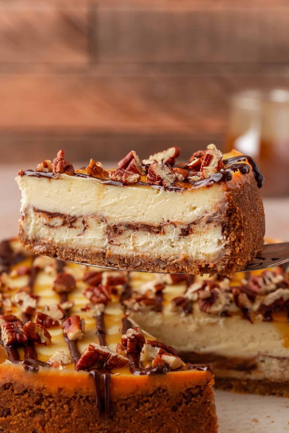 A close-up of a slice of cheesecake with a graham cracker crust, chocolate layer, and chopped pecans on top. The slice is being lifted from the whole cake in the background.