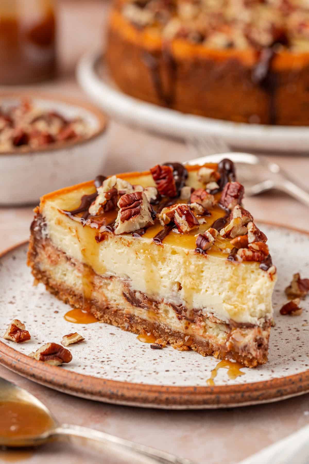 A slice of cheesecake topped with chopped pecans and caramel drizzle sits on a speckled plate, with more pecans and a whole cheesecake visible in the background.