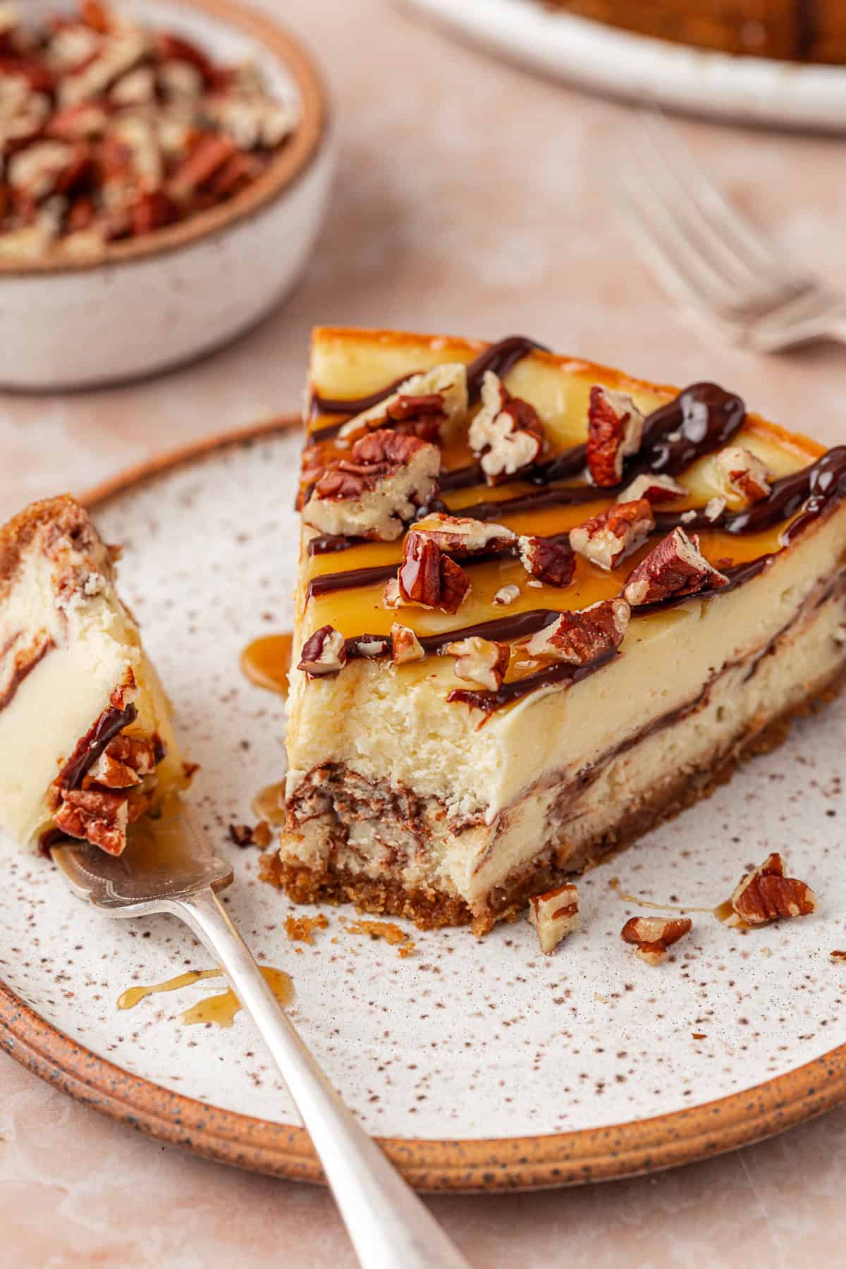 A slice of creamy cheesecake topped with chopped pecans, caramel, and chocolate drizzle sits on a speckled plate. A fork holds a bite of the cheesecake, and more dessert is in the background.