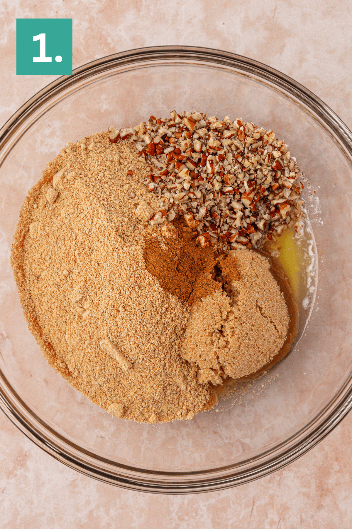 A glass bowl filled with graham cracker crumbs, chopped pecans, brown sugar, cinnamon, and melted butter, viewed from above. A green box with the number 1 is in the top left corner.