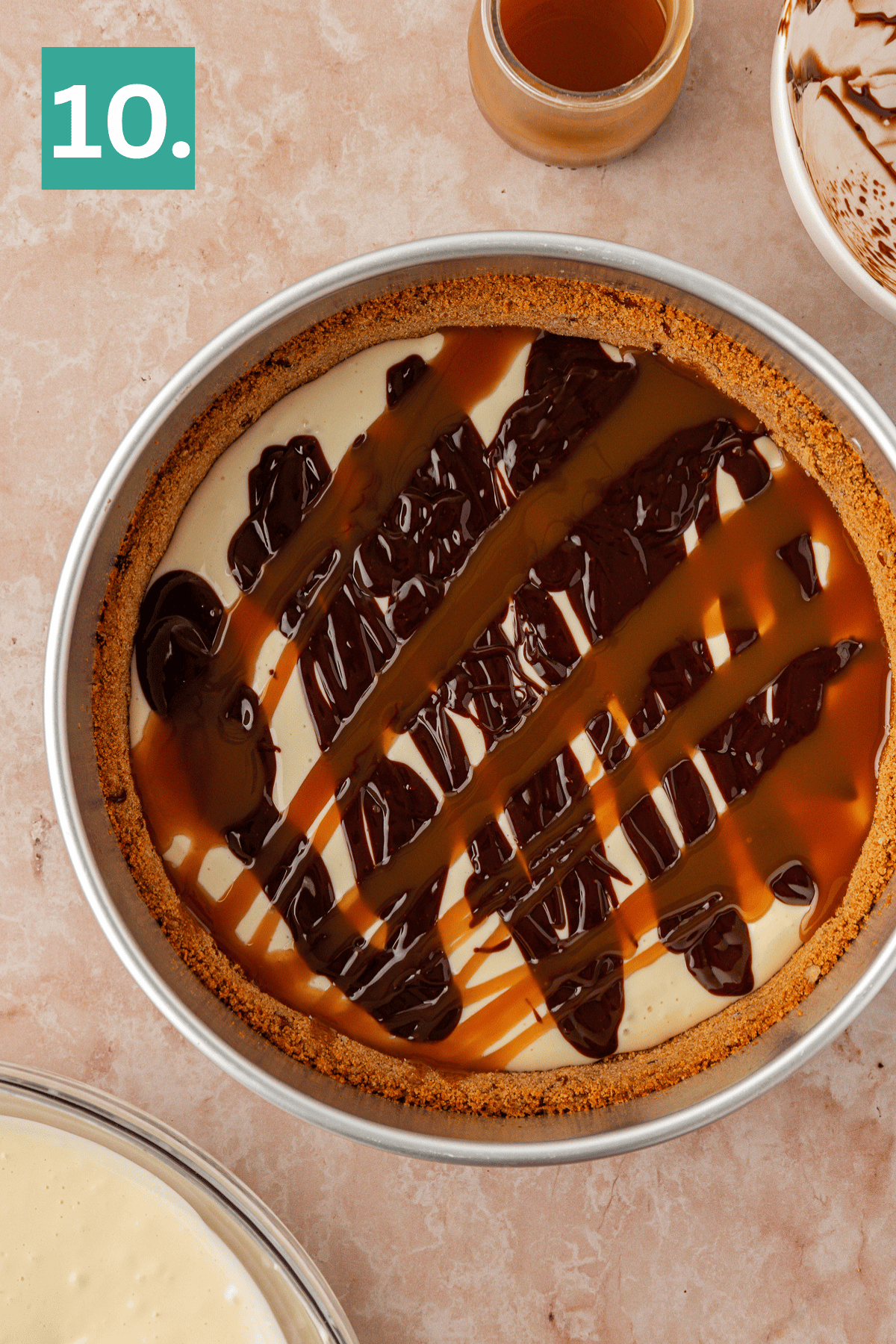 A round cheesecake in a pan is topped with drizzles of chocolate and caramel sauce. A jar of caramel and a bowl with chocolate remnants are nearby. A green box with the number 10 is in the top left corner.