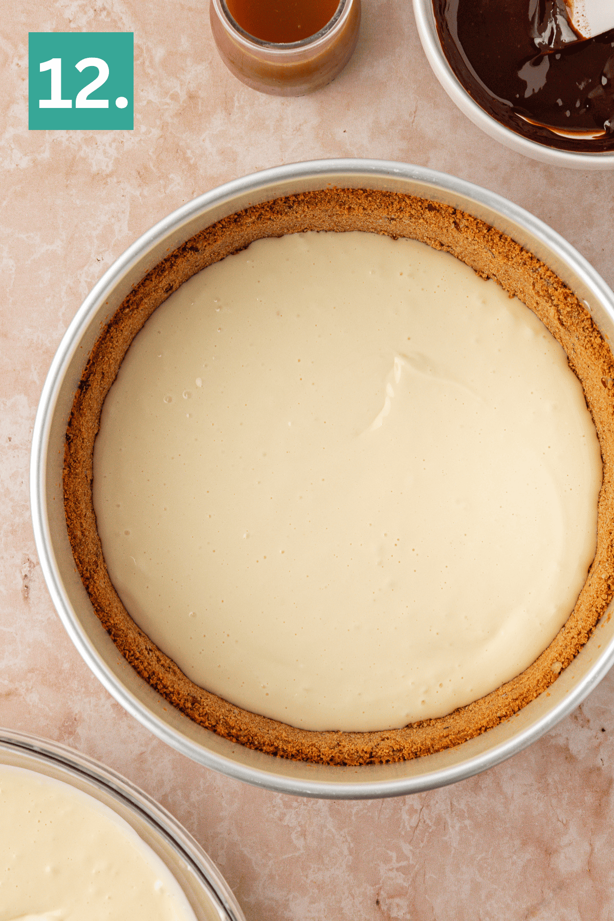 A cheesecake in a round pan with a graham cracker crust, unbaked and smooth on top, sits on a light countertop. A small bowl of caramel and a bowl of chocolate are nearby. A green box with the number 12 is in the corner.