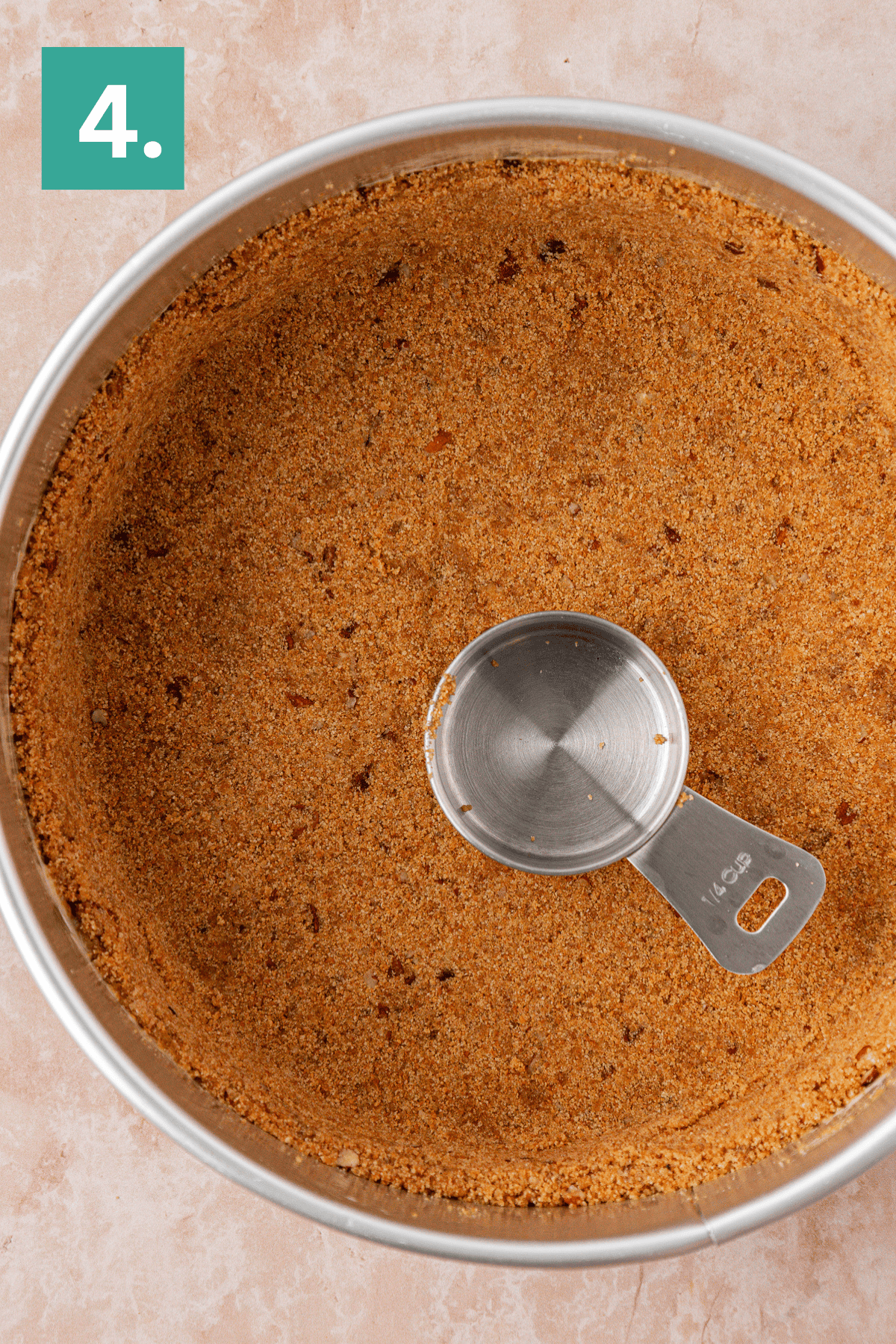 A round metal pan filled with an even layer of crumb crust and a metal measuring cup pressing the crust. A green box with the number 4 is in the top left corner.