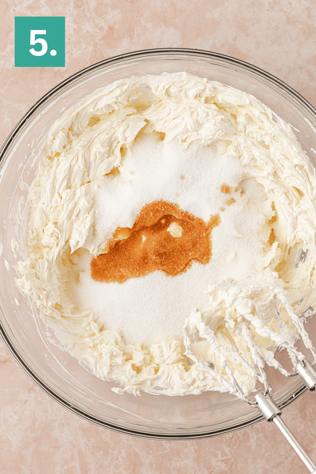 A glass mixing bowl with whipped cream cheese, sugar, and vanilla extract, shown with electric mixer beaters. A green square with the number 5 appears in the top left corner.
