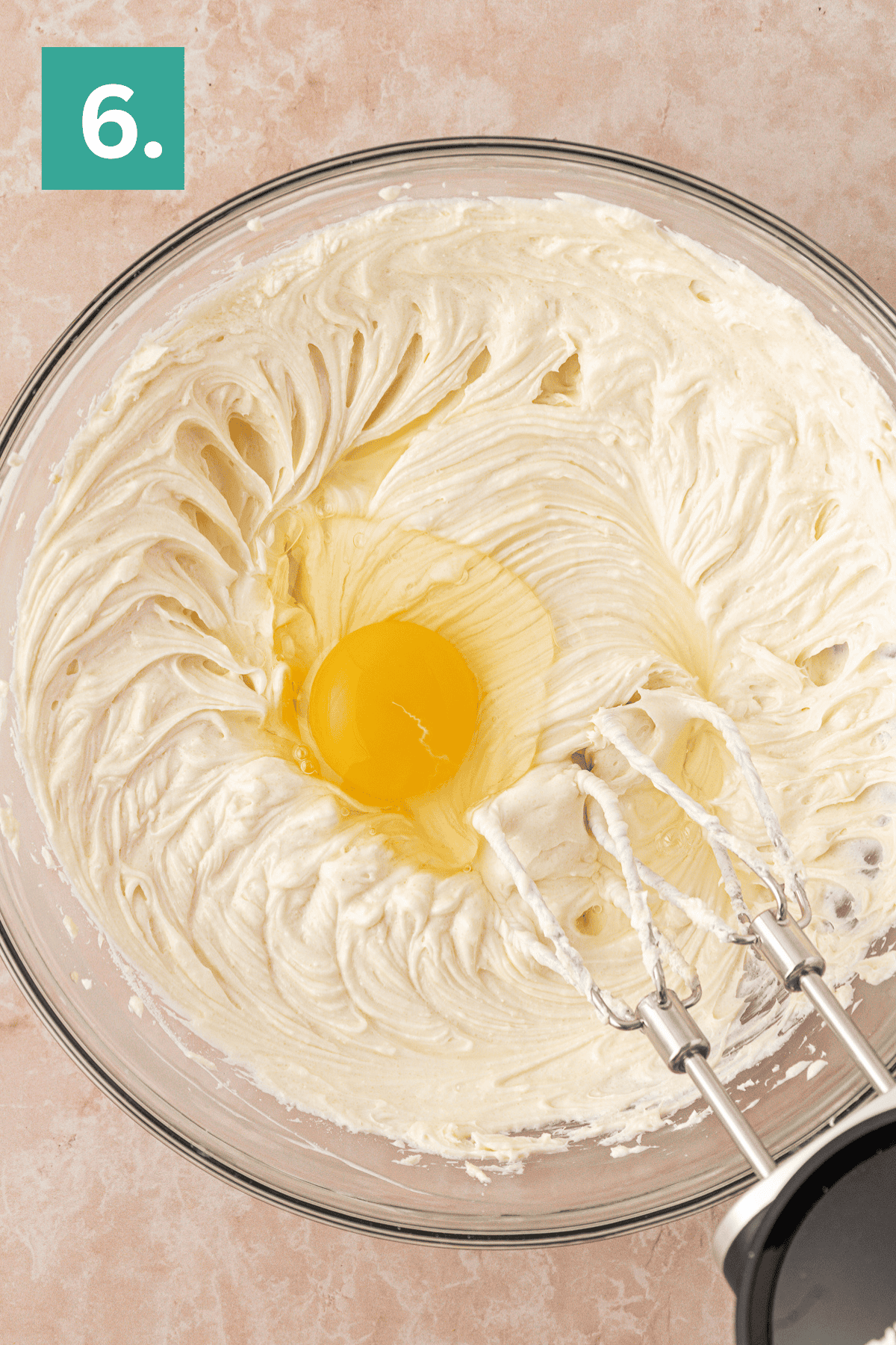 A glass bowl with creamy batter and a whole egg in the center, next to an electric hand mixer with beaters. A green box with the number 6 is in the top left corner.