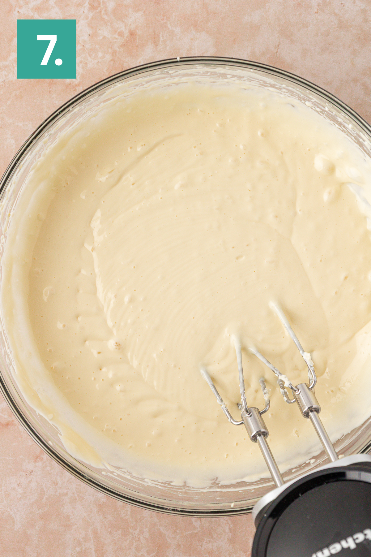 A glass bowl filled with smooth cake batter being mixed by an electric hand mixer, viewed from above. A green square with the number 7 is in the top left corner.
