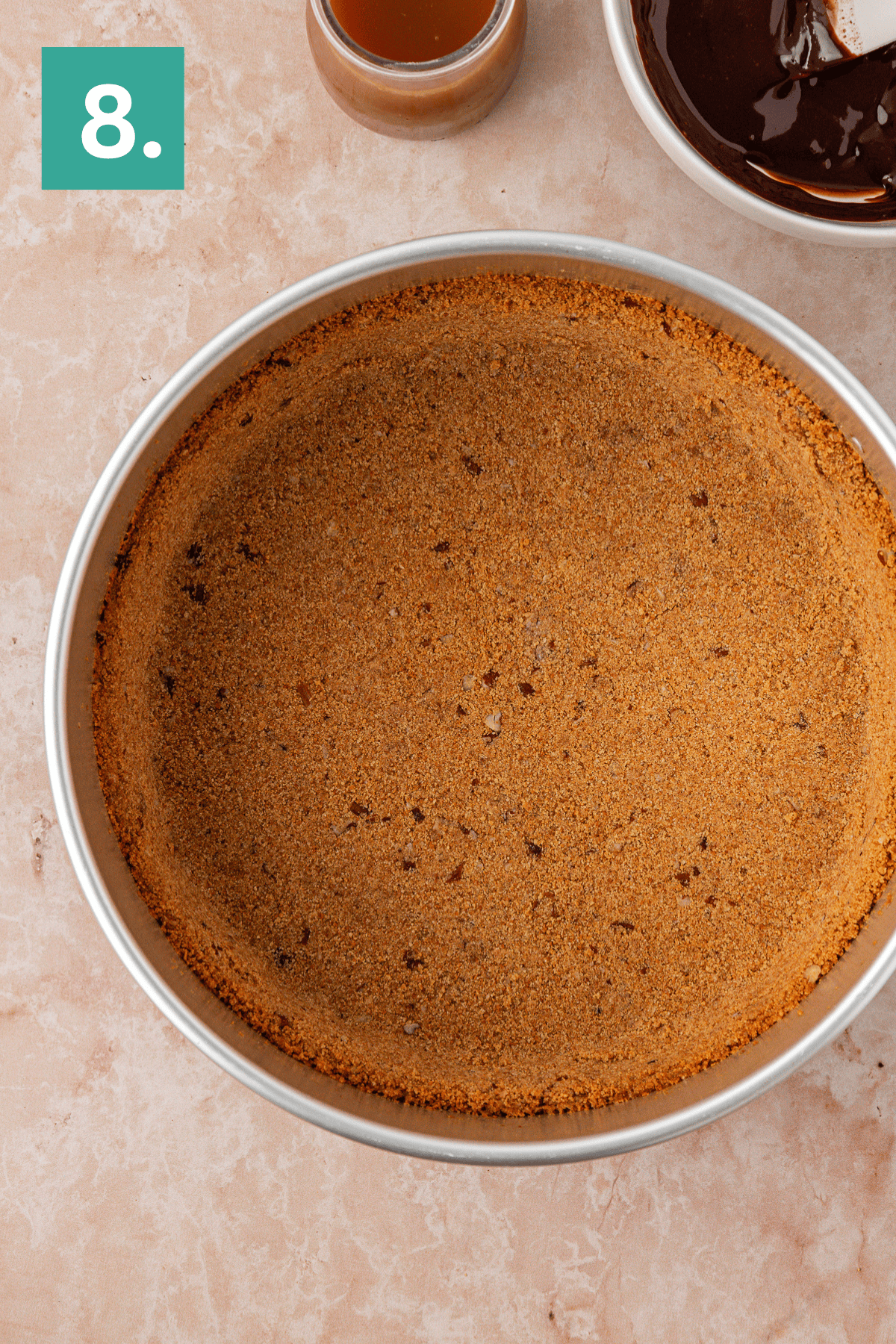 A round metal pie pan filled with a pressed graham cracker crust sits on a marble surface. The corner shows a bowl of melted chocolate, a small cup, and a green square with the number 8 in white.