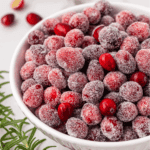 A white bowl filled with frosted, sugar-coated cranberries, with a few fresh cranberries mixed in. Green rosemary sprigs are visible at the edge of the image.