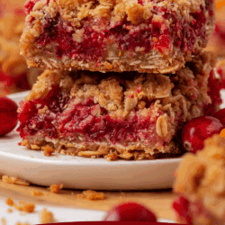 Two thick cranberry crumb bars stacked on a white plate, showing layers of oats and vibrant red cranberries. The background features whole cranberries and blurred crumb bar pieces. Text reads: CRANBERRY CRUMB BARS.
