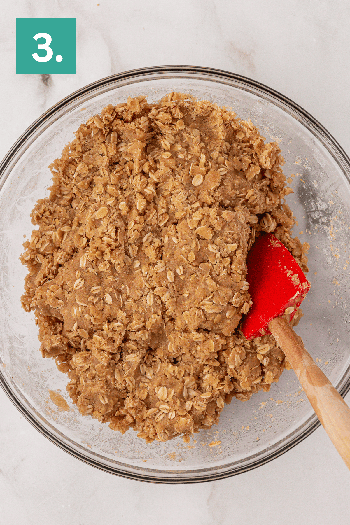 A glass bowl filled with oatmeal cookie dough is being mixed with a red spatula. A green box with the number 3 is in the top left corner. The bowl is placed on a light marble surface.