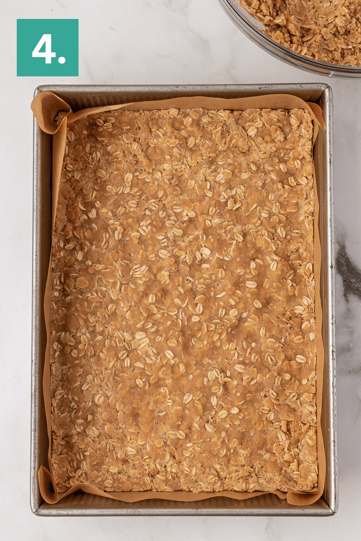 A square baking pan lined with parchment paper is filled with an unbaked oat mixture. A green box with the number 4 is in the top left corner. The pan sits on a white marble surface.