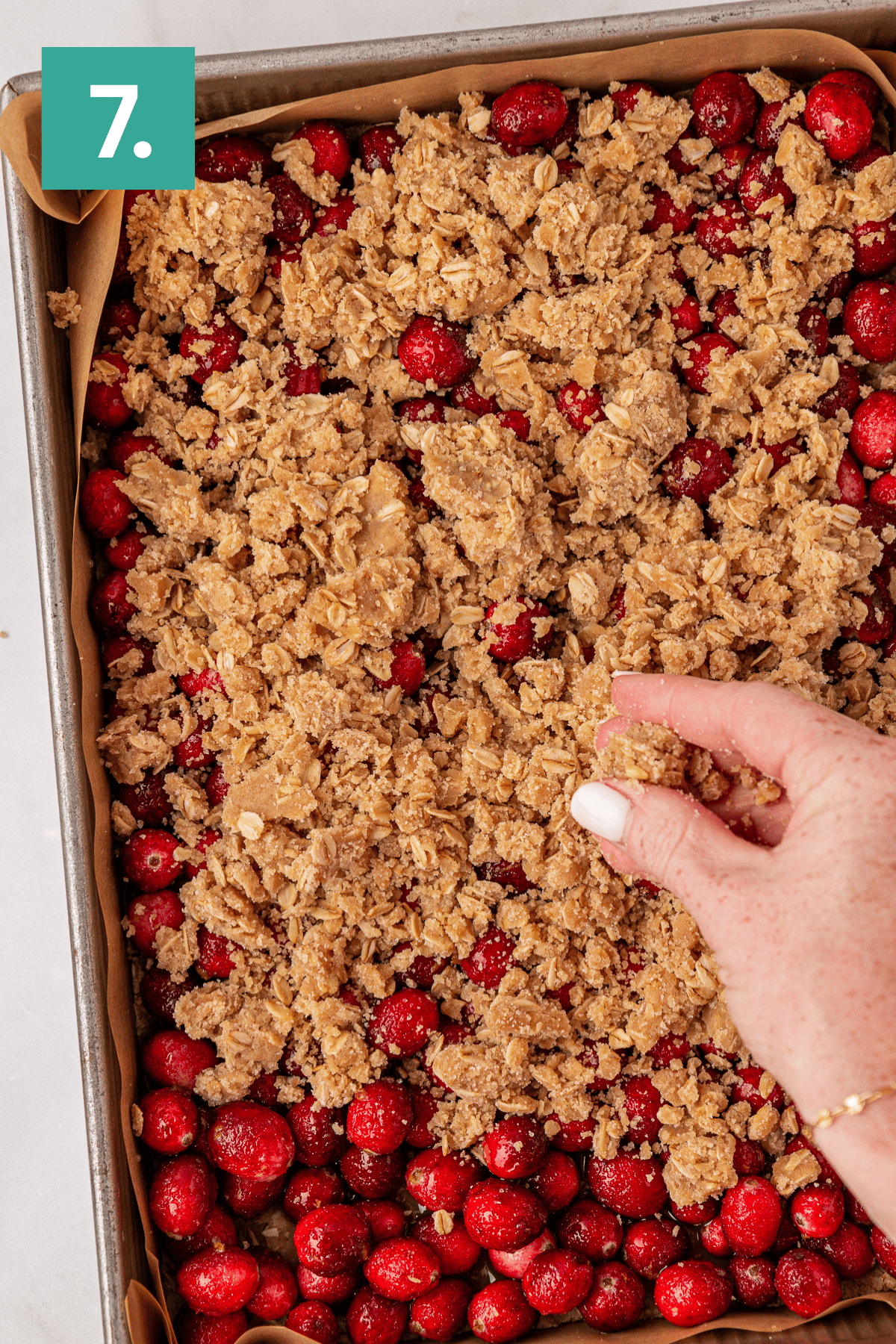 A hand sprinkles crumbly oat topping over a baking sheet filled with fresh cranberries. A green box with the number 7 is in the top left corner.
