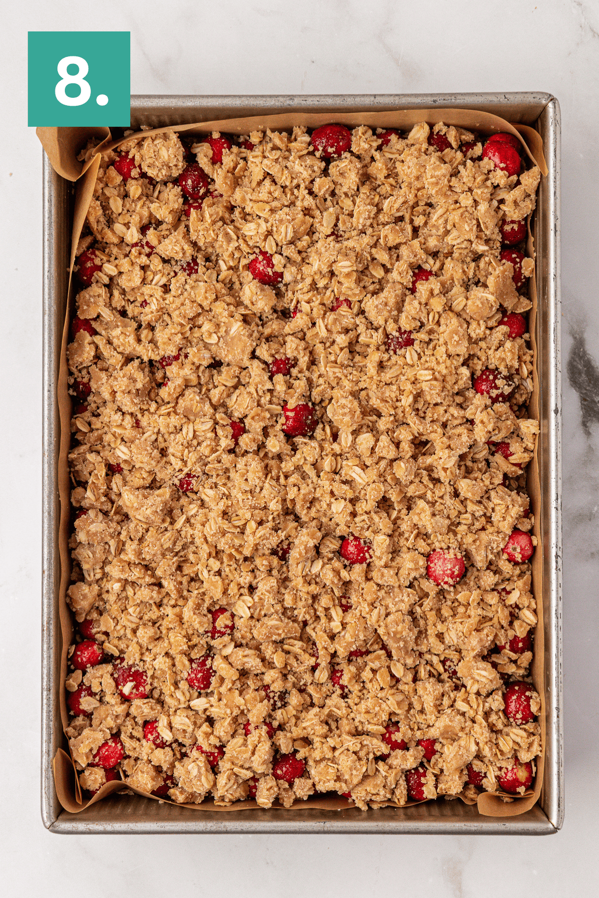 A rectangular baking pan lined with parchment paper is filled with an unbaked cranberry crumble, topped with a golden oat and brown sugar streusel. A green square with the number 8 is in the top left corner.