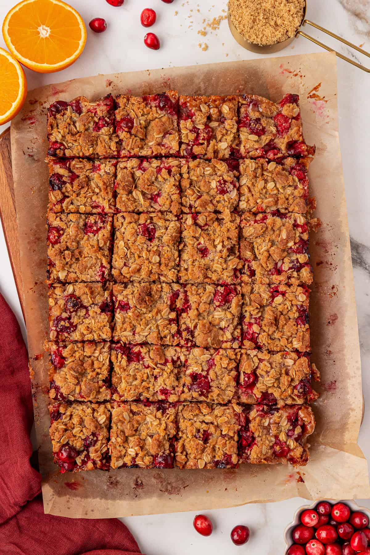 A tray of oat and cranberry bars cut into squares on parchment paper, surrounded by fresh cranberries, sliced oranges, and a measuring cup of brown sugar on a marble surface.