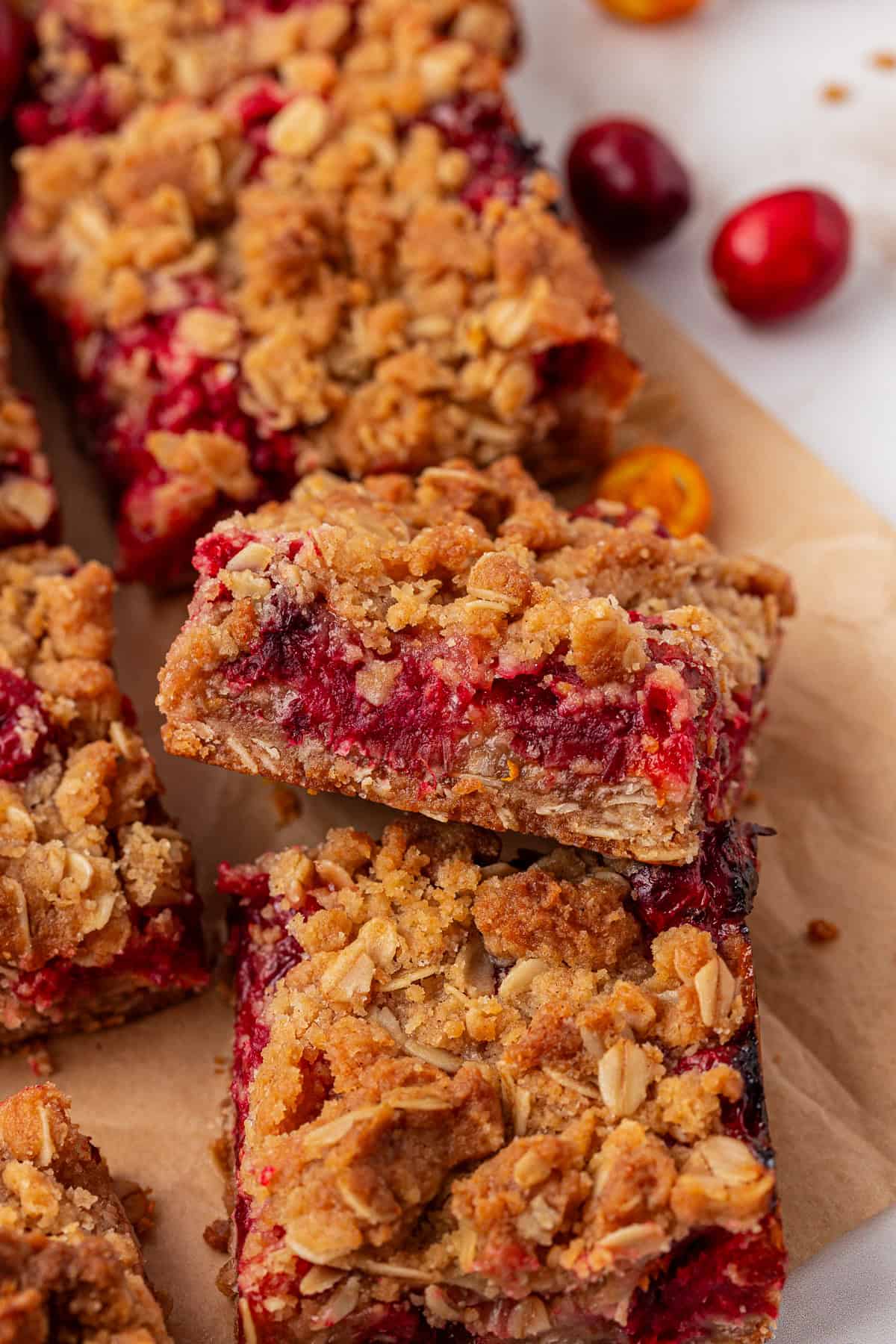 Close-up of oat crumble bars filled with a red fruit, likely cranberry filling. The bars have a golden, crumbly oat topping and are stacked on parchment paper, with a few whole cranberries nearby.