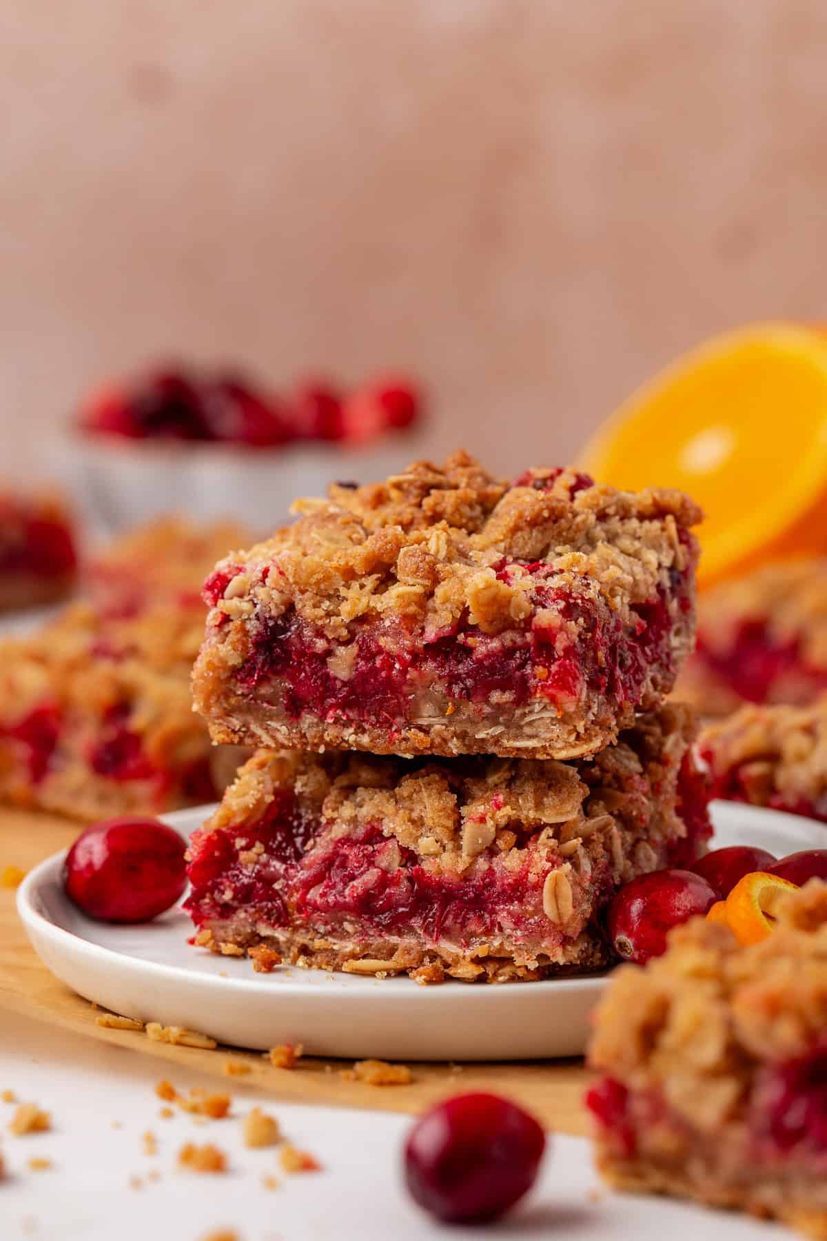 two cranberry bars stacked on a small white plate with scattered whole cranberries around them and more bars in the background