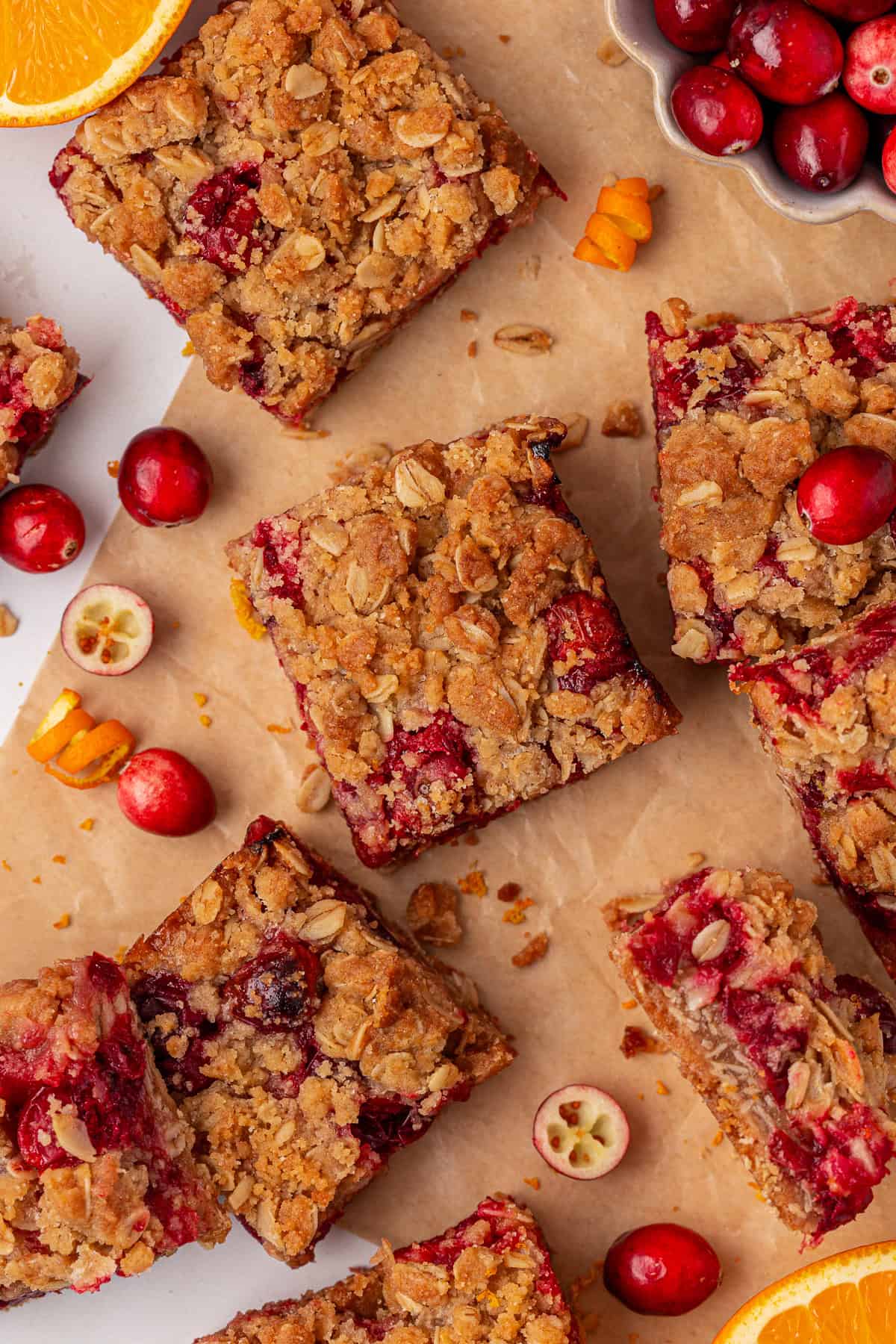 Cranberry oat crumble bars are arranged on brown parchment paper, surrounded by fresh cranberries and orange slices. The bars have a golden oat topping and visible red cranberries throughout.