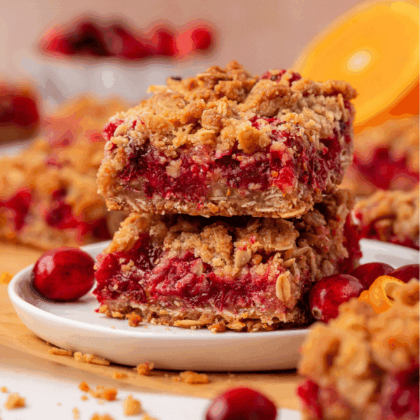Two crumbly fruit bars with a red filling are stacked on a white plate, surrounded by fresh cranberries and orange slices. More bars and ingredients are blurred in the background.