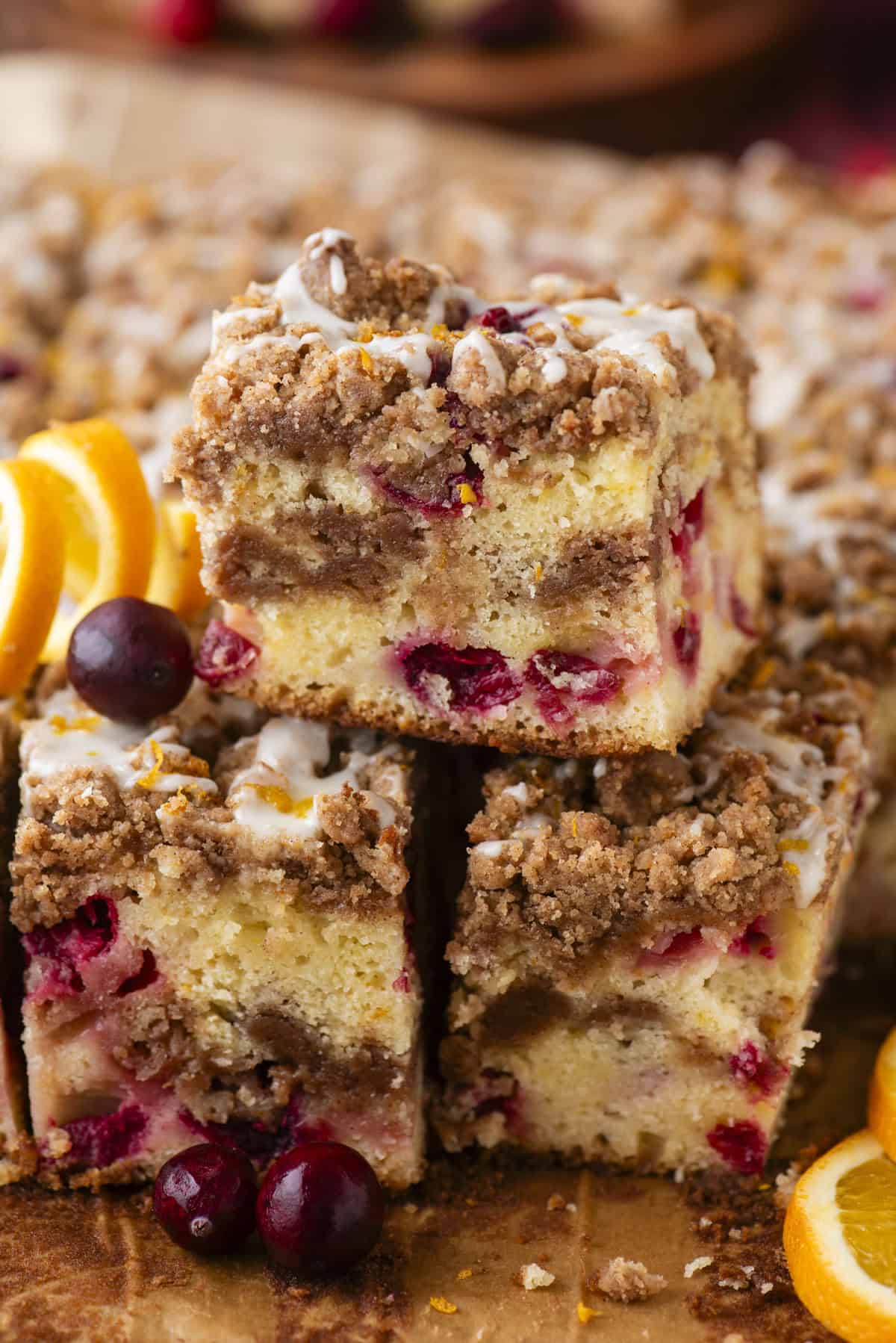 A close-up of three squares of cranberry coffee cake with cinnamon streusel topping and a light glaze. Fresh cranberries and orange slices are arranged around the cake pieces.