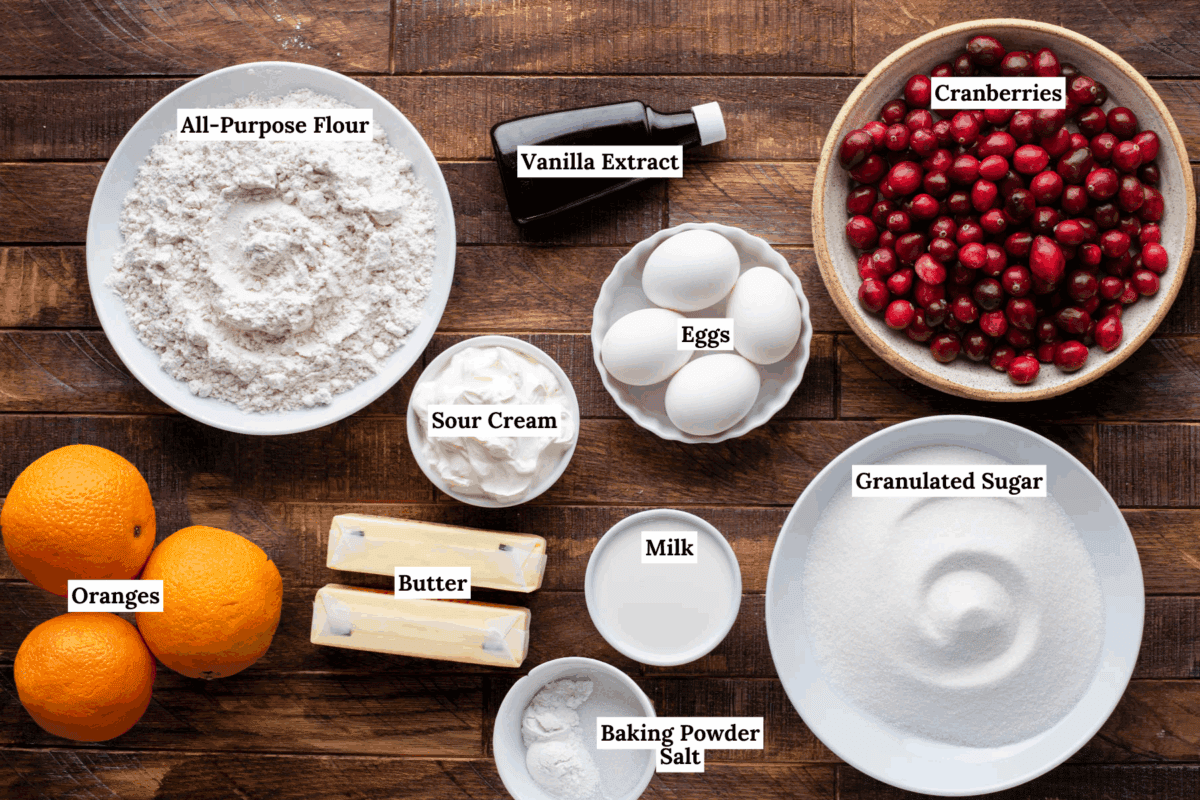 A top-down view of baking ingredients on a wooden table, including flour, eggs, cranberries, sugar, oranges, sour cream, butter, milk, baking powder, salt, and vanilla extract, each labeled.