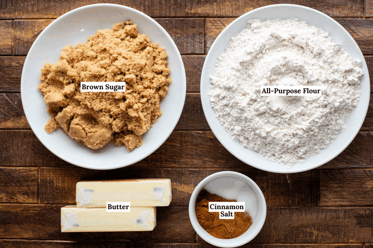 Four baking ingredients on a wooden surface: a bowl of brown sugar, a bowl of all-purpose flour, two sticks of butter, and a small bowl with cinnamon and salt. Each item is labeled with text.