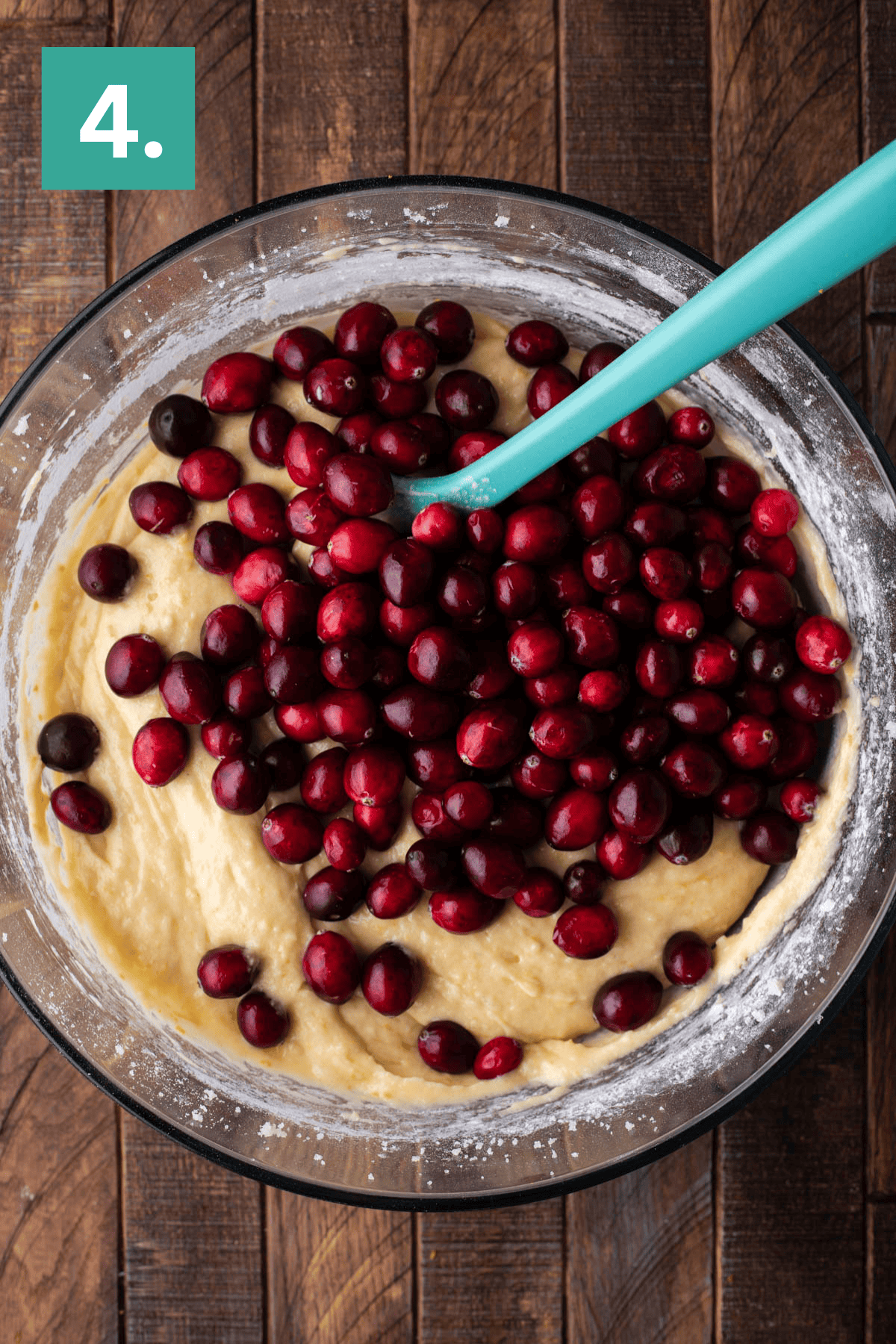 A glass bowl filled with cake batter and fresh cranberries being mixed with a teal spatula, sitting on a wooden surface. A green square with the number 4 is in the top left corner.