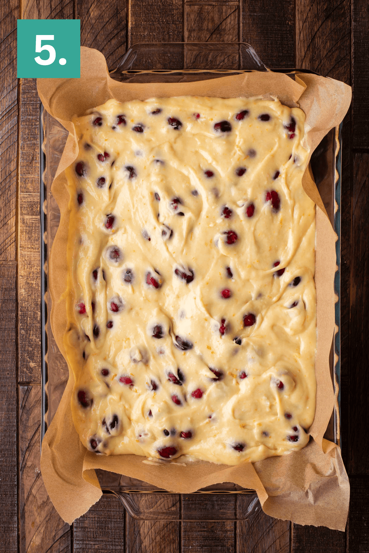 A glass baking dish lined with parchment paper and filled with raw batter dotted with red and dark berries sits on a wooden surface. A green square with the number 5 is in the top left corner.