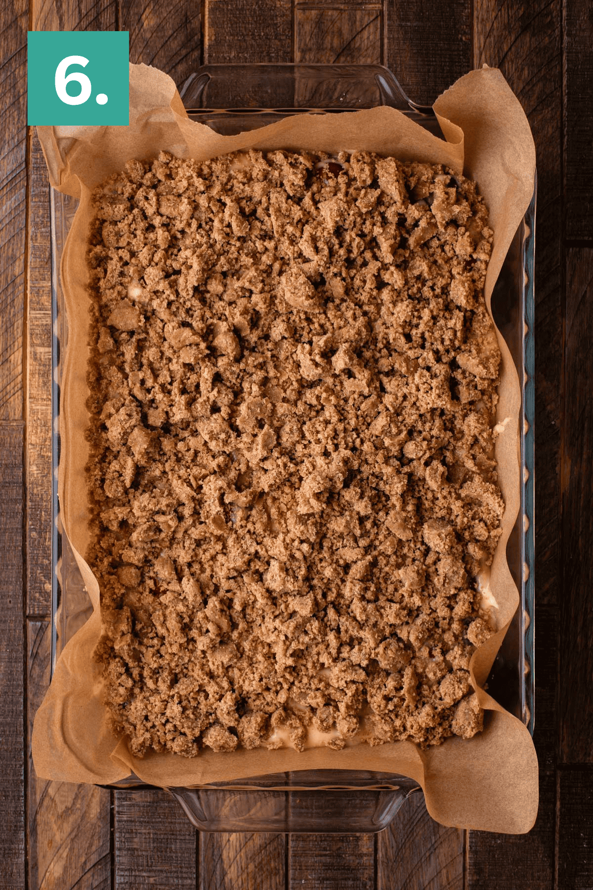A glass baking dish lined with parchment paper holds a large crumb-topped dessert, likely coffee cake or crumble, on a wooden surface. A green box with the number 6 appears in the top left corner.