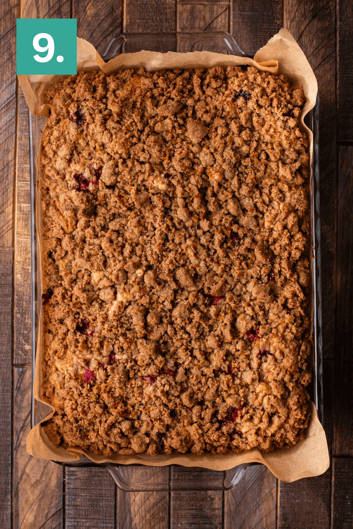 A large rectangular crumb cake with a golden-brown streusel topping sits in a glass baking dish lined with parchment paper on a wooden surface. A green box with the number 9 is in the top left corner.