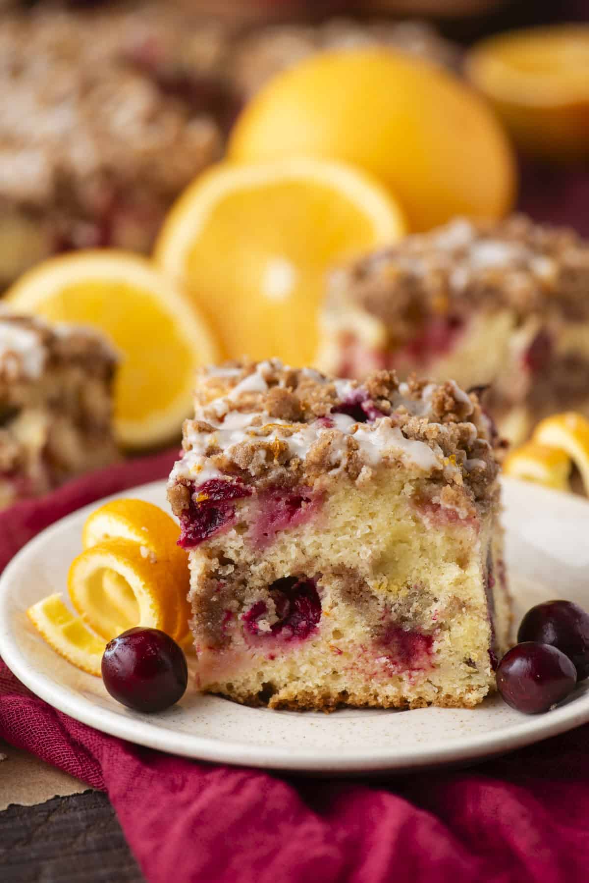 A slice of crumb-topped coffee cake with cranberries and icing sits on a plate, garnished with fresh orange slices and whole cranberries. Whole oranges and more cake are blurred in the background.