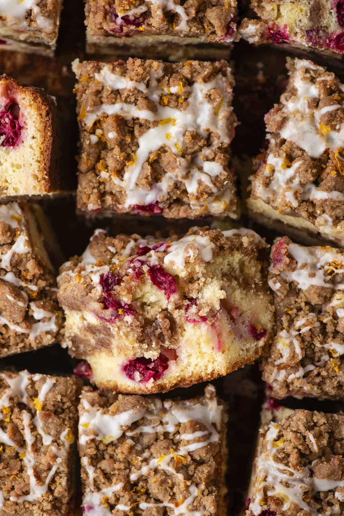 Close-up of sliced coffee cake squares topped with a crumbly streusel, visible raspberries, and a drizzle of icing. The cake is golden and looks moist with bits of fruit and crunchy topping.