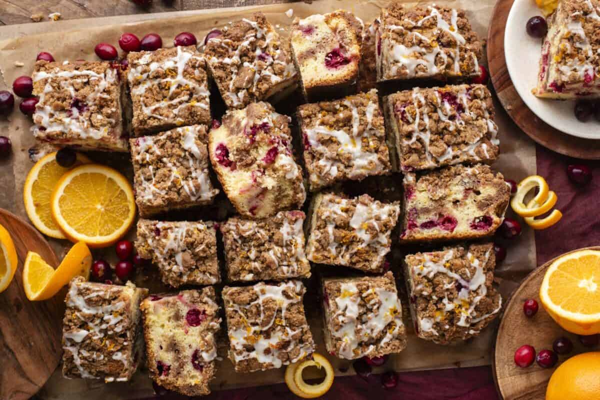 Overhead view of sliced coffee cake squares with cranberries and orange zest, topped with drizzled icing and streusel. Fresh orange slices, cranberries, and orange peels are scattered around on a wooden surface.